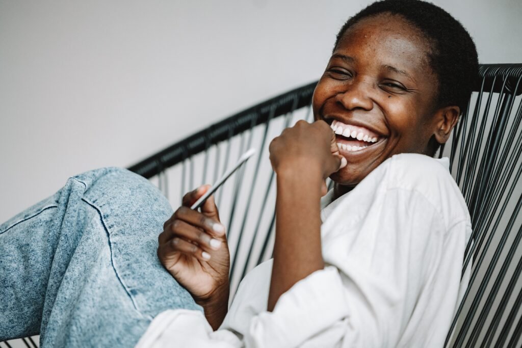 A cheerful African woman laughs while relaxing on a comfortable chair indoors.